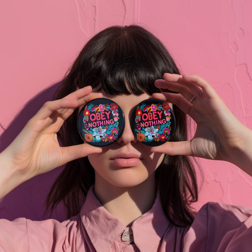 A black round pinback button featuring floral embroidery-style designs and bold red text that reads "Obey Nothing."